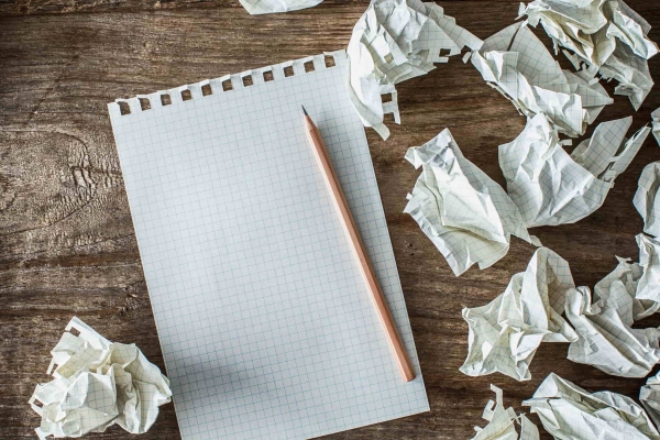 A blank sheet of rectangular paper with a pencil on a wooden background with scrunched up pieces of paper surrounding it