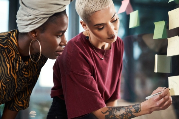 On the left of the photo, a Black woman wearing a grey head wrap looks on as her colleague - stood on the right a white woman with cropped, bleached hair and tattooed arm - writes on a sticky-note stuck to a glass wall. Both are bending down.