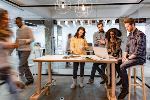 Two white men, a black woman and a white woman sit around a table looking at a laptop while other figures are blurred in the bacground walking past the glass office in which they are sat or stood