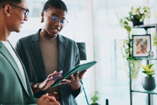 In a modern office two Black male and female colleagues stand looking at a tablet. Both wear dark green smart clothing, hold a green iPad and are surrounded by plants on shelves