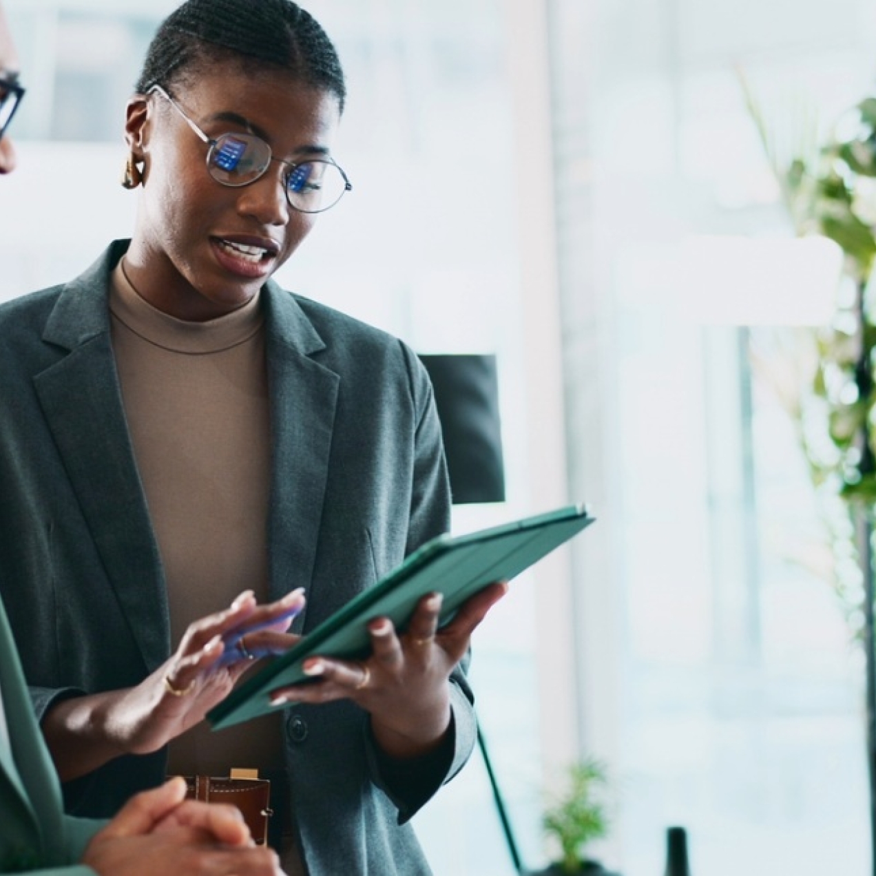 In a modern office two Black male and female colleagues stand looking at a tablet. Both wear dark green smart clothing, hold a green iPad and are surrounded by plants on shelves