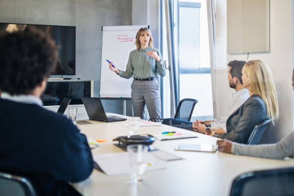 A white woman stands while addressing colleagues of different genders and ethnicities sat around a board table. Next to her is a display board with the words crisis management