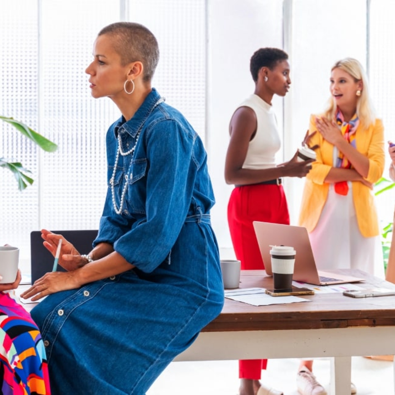 Six business women of different ages and ethnicities. Two in the foreground are perched on a table. The others are stood chatting behind. All are wearing bright and colourful clothes.