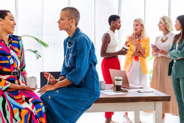 Six business women of different ages and ethnicities. Two in the foreground are perched on a table. The others are stood chatting behind. All are wearing bright and colourful clothes.