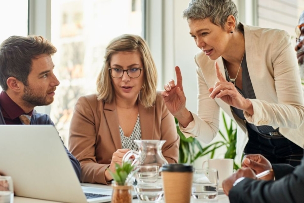 Huddled around a laptop, a white woman with short grey hair stands with her fingers raised while three colleagues listen - a younger white man with brown hair, a younger white woman and a Black man with grey beard - look on.