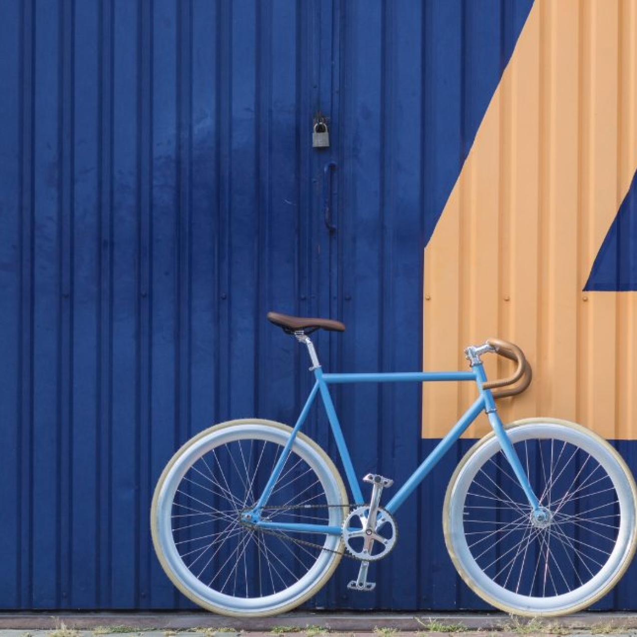 A turquoise bike with white wheel trims stood against blue corrugated doors which feature a yellow painted four numeral. To the right, a white man wearing denim stands side on looking at the bike.