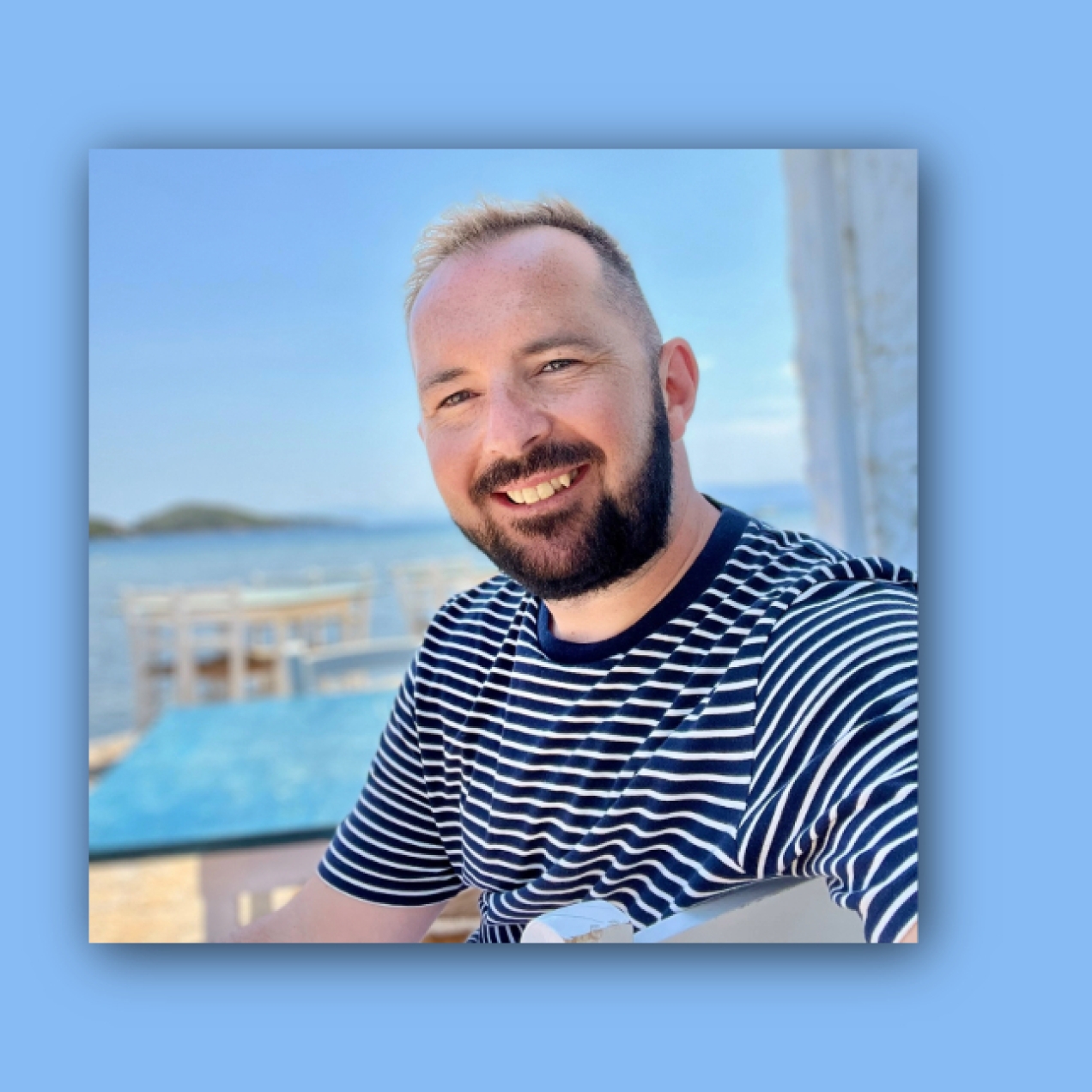 On a blue background, a square colour portrait of Paul Quinn. Paul is a white man with a dark beard wearing a striped top.