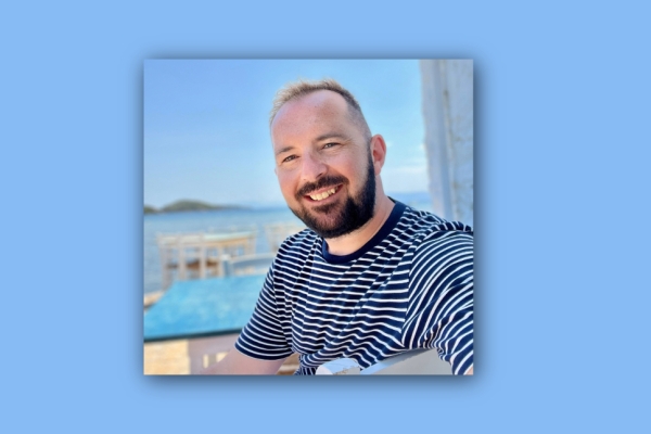 On a blue background, a square colour portrait of Paul Quinn. Paul is a white man with a dark beard wearing a striped top.