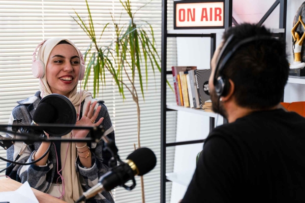 A smiling woman wearing large headphones over her cream hijab engages in conversation during a podcast. She faces an unidentified man with dark hair and black shirt. They are surrounded by microphones, laptop, notepad and an on-air sign upon a shelf.