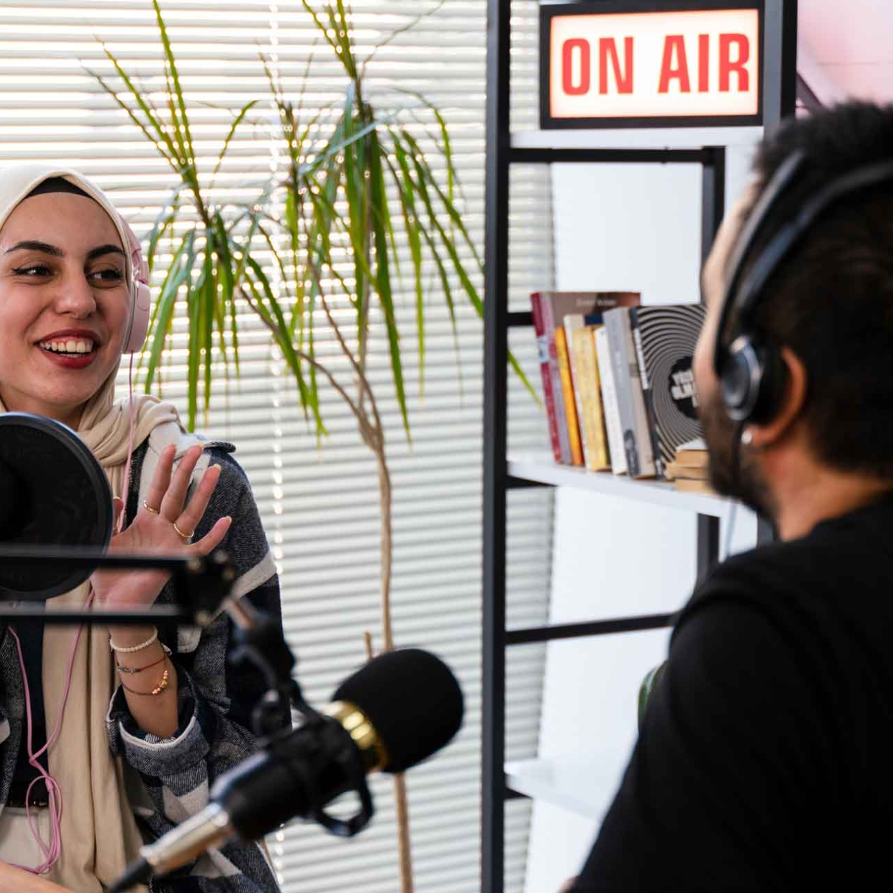 A smiling woman wearing large headphones over her cream hijab engages in conversation during a podcast. She faces an unidentified man with dark hair and black shirt. They are surrounded by microphones, laptop, notepad and an on-air sign upon a shelf.