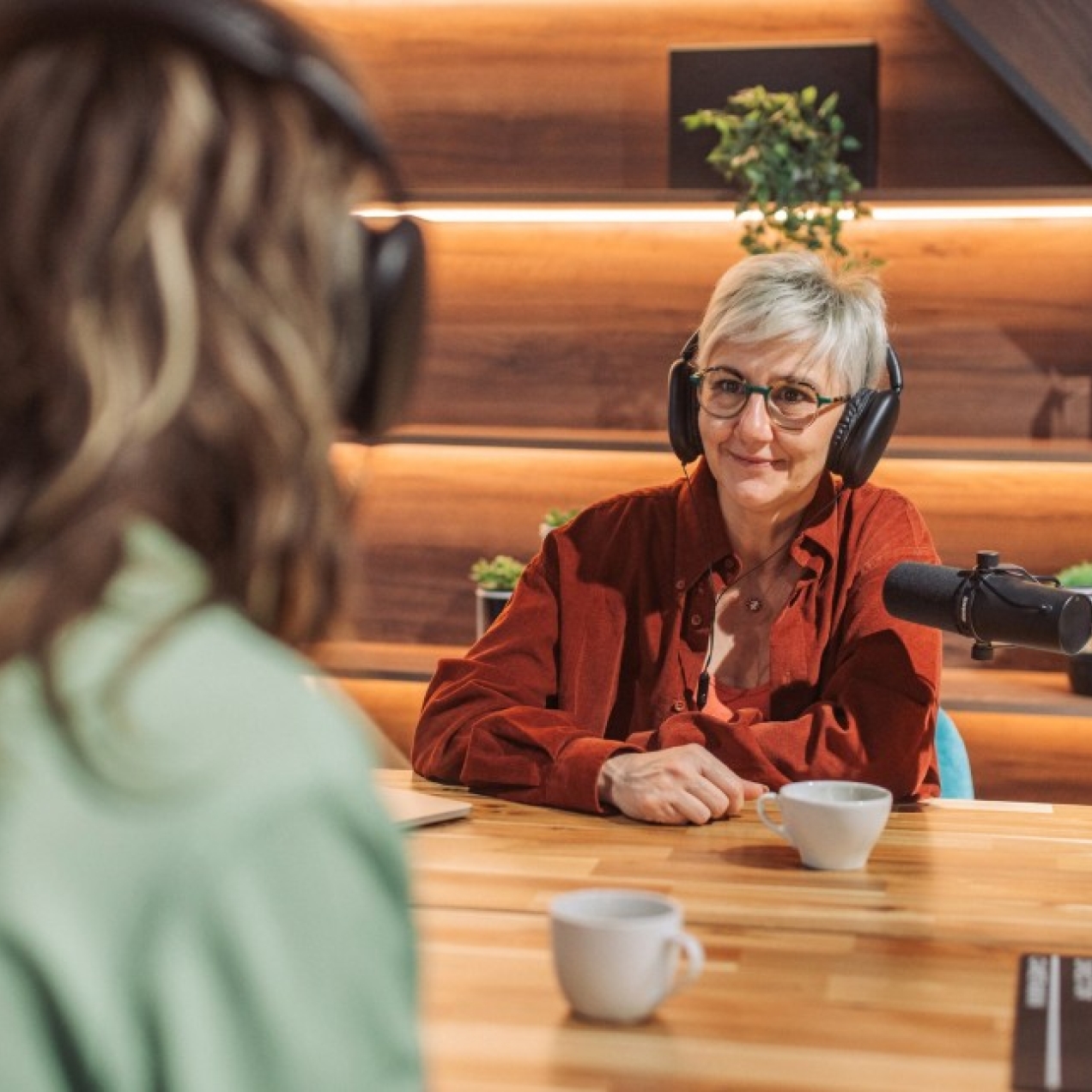 Within the eaves of wood panelled room an older white woman with short grey hair and wearing headphones is in discussion across a microphone with another unidentifiable woman whose back faces the camera