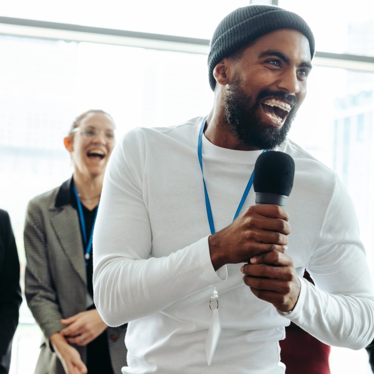 A smiling Black man wearing a dark beanie hat and white long sleeve top and a blue lanyard holds a microphone. A few women are stood smiling behind him in front of a window.