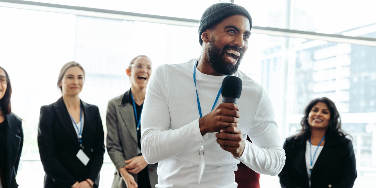 A smiling Black man wearing a dark beanie hat and white long sleeve top and a blue lanyard holds a microphone. A few women are stood smiling behind him in front of a window.