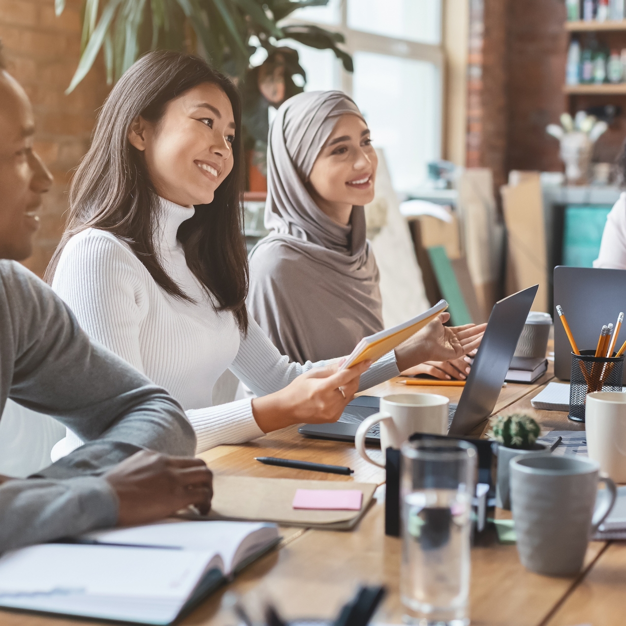 Smiling colleagues sat around a table with books, mugs and laptops. From left to right: a young Black man with short hair and grey jumper; a south-east Asian woman with shoulder length hair and white jumper; an Asian woman wearing a grey hijab; a Bla