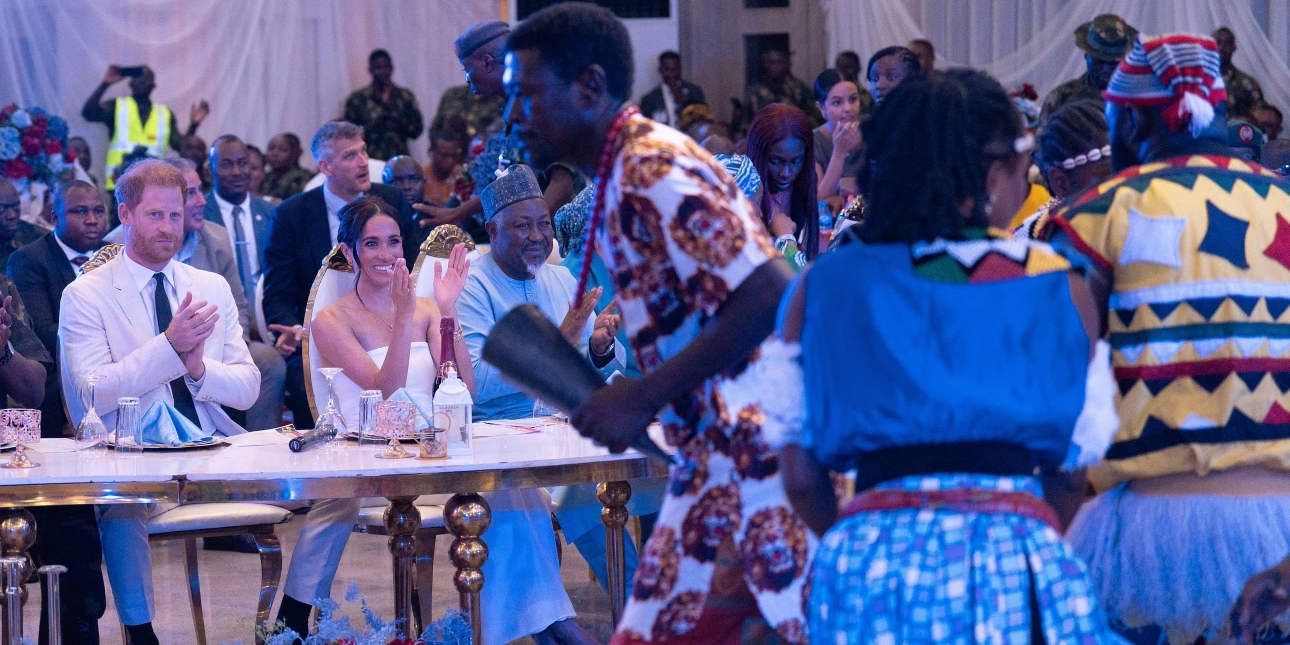 A smiling Prince Harry and Meghan surrounded by delegates at a table, clap at dancers in front of them