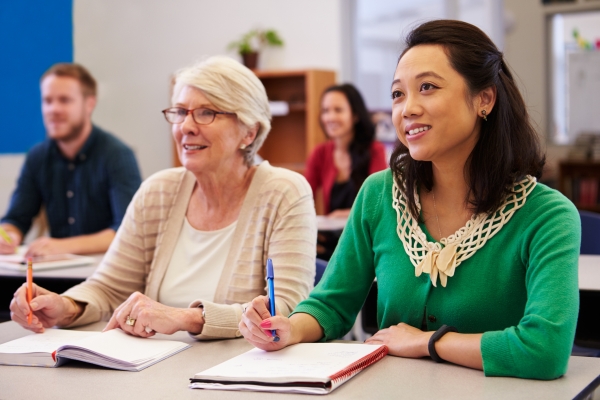 Two women sat at a desk with open notebooks in a classroom. The spectacle-wearing woman on the left is older, white, with short grey hair. The younger woman on the right is Asian, with shoulder length dark hair and wears a green top. Out of focus beh