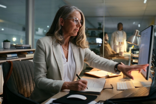 In a dimly lit office a white middle-aged woman with shoulder-length grey hair points at a computer screen with her left hand while writing with her other hand. She is wearing a grey blazer.