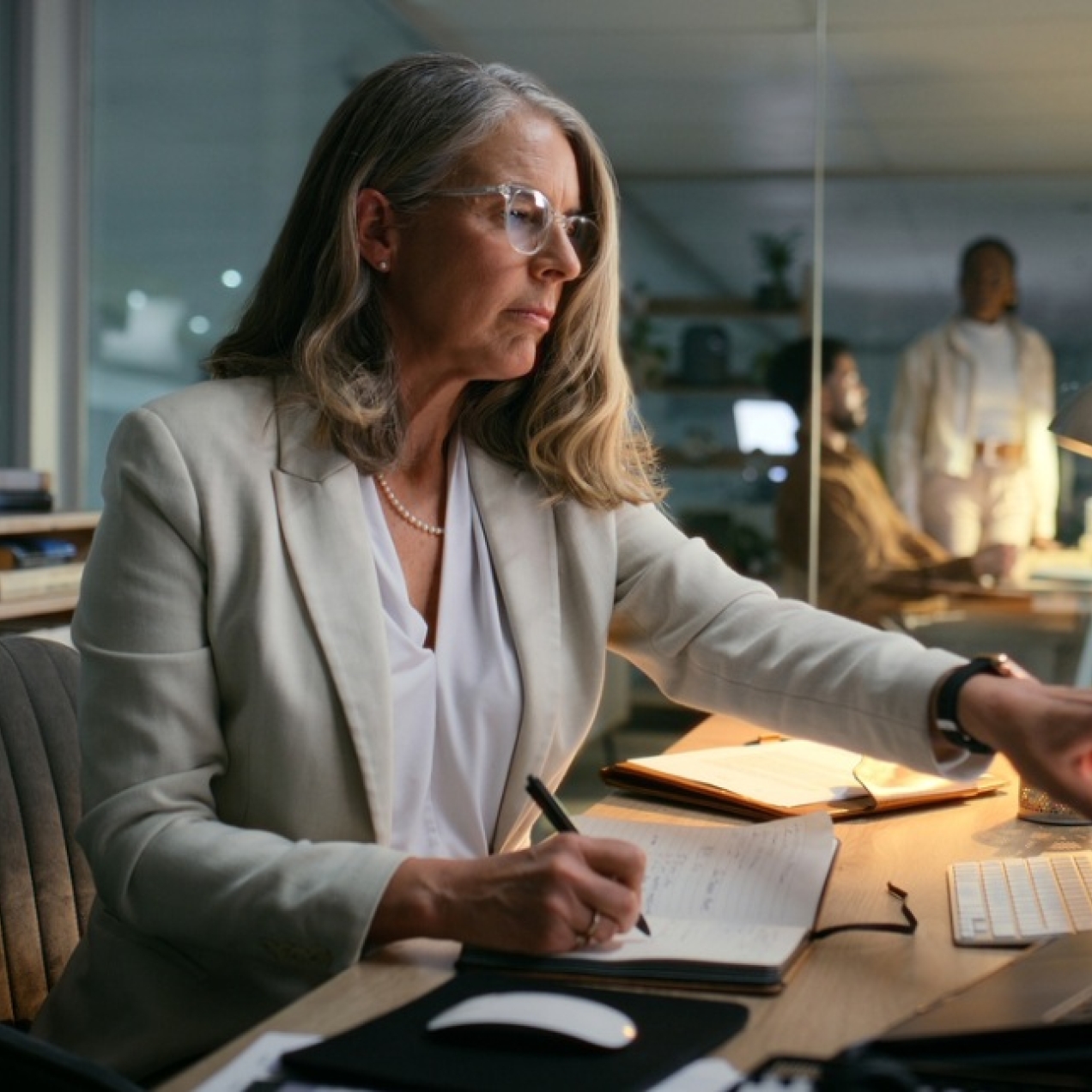 In a dimly lit office a white middle-aged woman with shoulder-length grey hair points at a computer screen with her left hand while writing with her other hand. She is wearing a grey blazer.