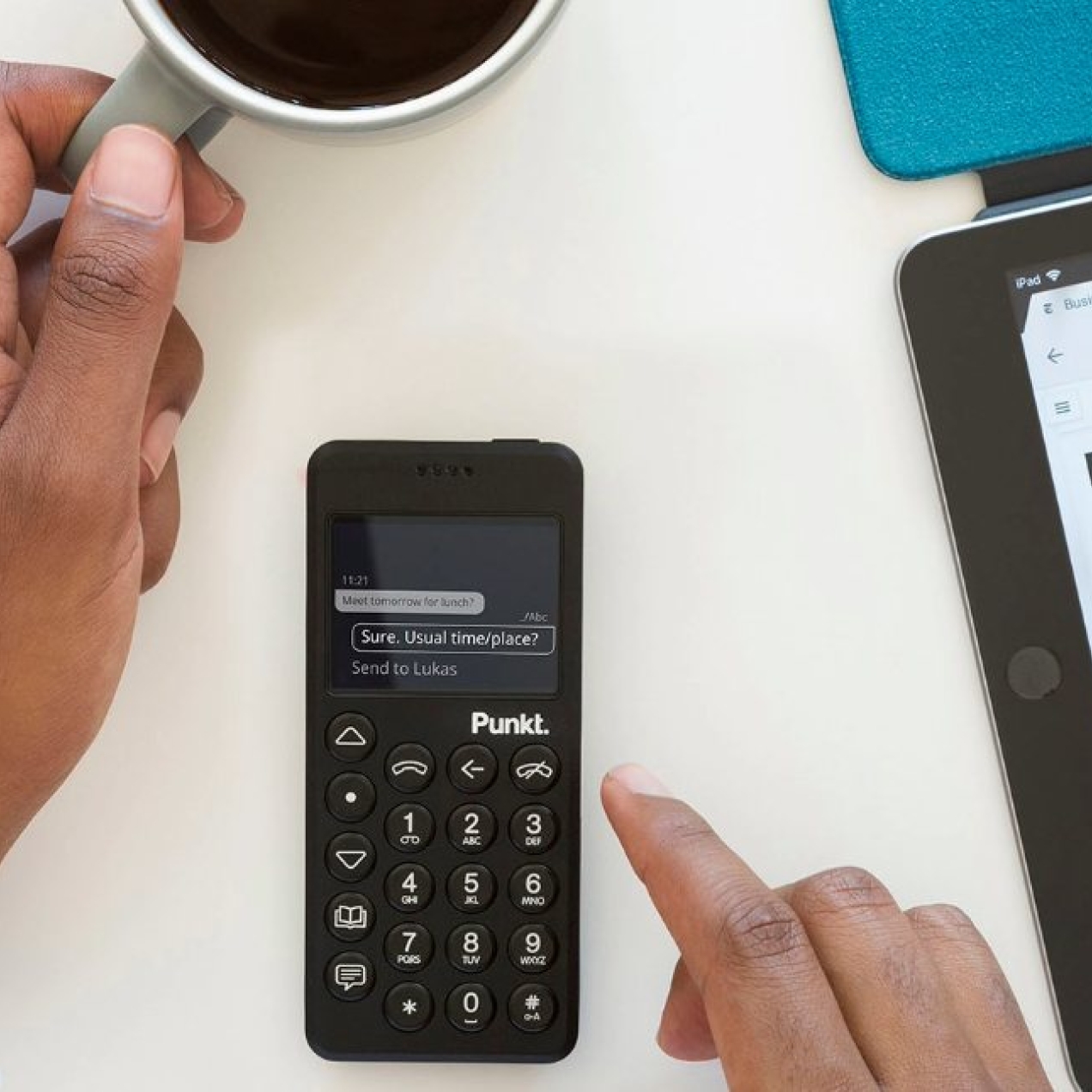An unidentified right hand operates a Punkt device while a cup of coffee is held in the left hand. Adjacent is a tablet with a screen showing a business day calendar.