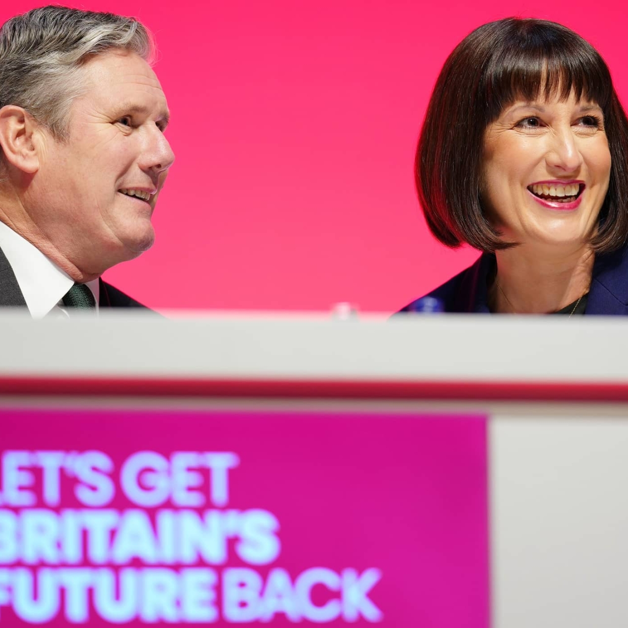 Keir Starmer, a white man with grey hair, and Rachel Reeves, a white woman with a dark brown bob, wear sat smiling at a podium. The background is red and the slogan beneath them reads let's get Britain's future back
