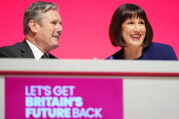 Keir Starmer, a white man with grey hair, and Rachel Reeves, a white woman with a dark brown bob, wear sat smiling at a podium. The background is red and the slogan beneath them reads let's get Britain's future back