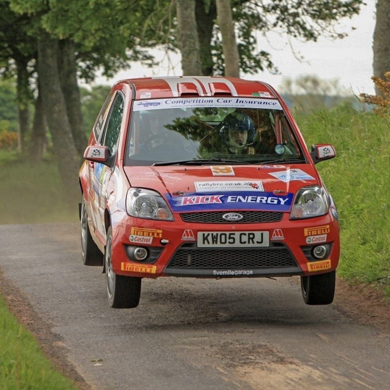 A red Ford Focus rally car emblazoned with sponsor logos takes to the air as it speeds down a narrow track with green verges either side
