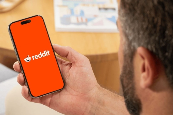 A white man holds a mobile phone featuring the reddit logo on an orange background on the screen. His head and dark hair is partially visible.