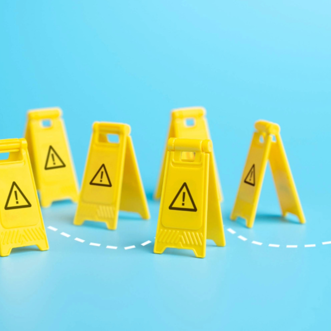 A computer generated image of seven yellow plastic A-boards with black printed exclamation marks on the front. They are placed in random position. A wavy, white dashed line denotes a path through them. The floor and background is bright blue.