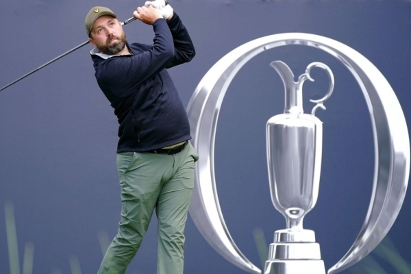 Golf influencer Rick Shiels tees off. He is a white man with a beard who wears a green cap, blue top and green trousers. He is in front of the a blue hoarding with a silver emblem.