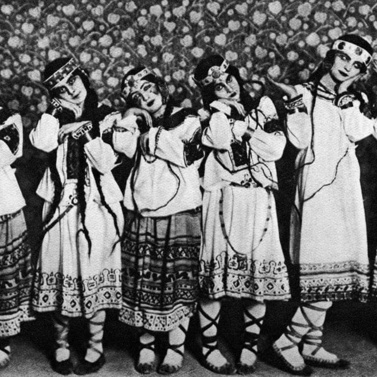 A black and white photograph of six dancers in embroidered tunics, long dresses and boots laying their heads on their hands