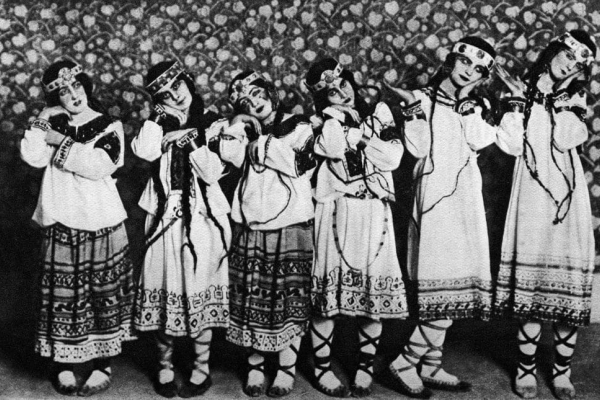 A black and white photograph of six dancers in embroidered tunics, long dresses and boots laying their heads on their hands