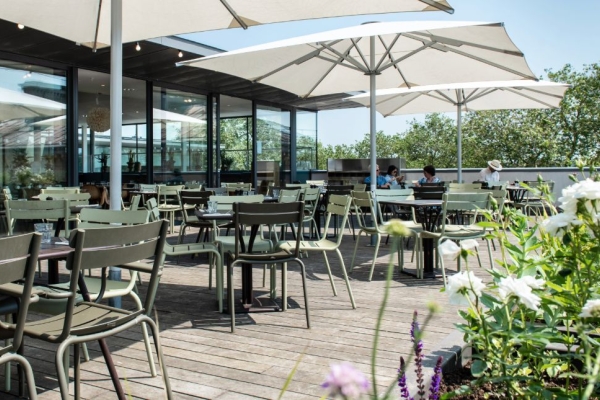 A restaurant roof terrace on a sunny day with green chairs, large white parasols, flower beds and a blue sky.