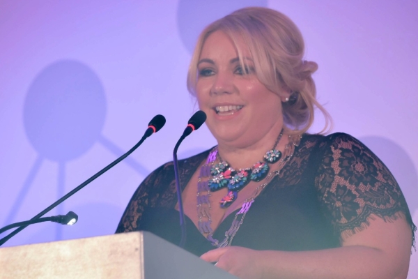 Samantha Livingstone stood at a lecturn under pink lighting. Samantha is a white woman with blond hair who is wearing a black top with laced sleeves.