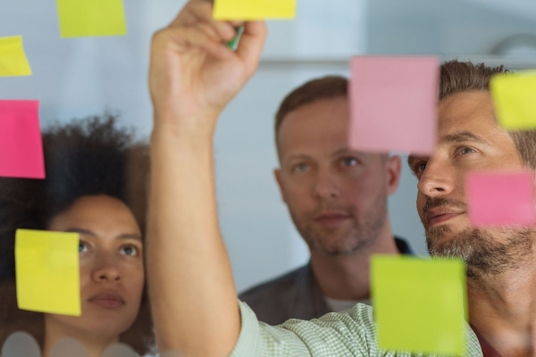 Looking through a glass wall adorned with multicoloured sticky notes at three colleagues - a Black woman and two white men. The man on the right is writing on one of the notes as the other look on.