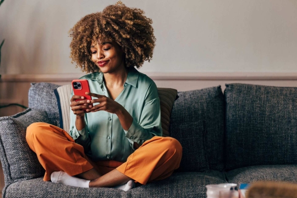 A smiling Black woman using her smartphone while sitting crossed legged on the sofa in a modern but cosy living room.