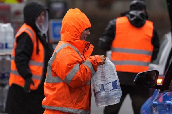 A worker in orange hi-vis clothing hands over bottled water to unseen recipients of a blue car.