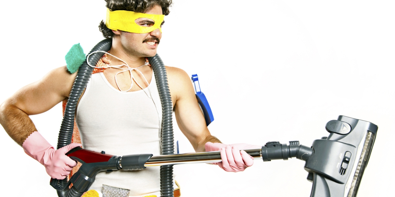 A white man with dark curly hair, moustache and white vest wears a yellow eye mask, pink rubber gloves and holds a vacuum attachment in his hands. Tied to his neck as a cape is an iron board cover. He has brushes and scourers attached to his belt.