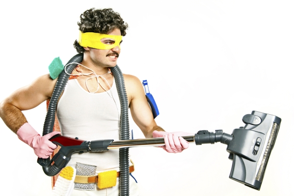 A white man with dark curly hair, moustache and white vest wears a yellow eye mask, pink rubber gloves and holds a vacuum attachment in his hands. Tied to his neck as a cape is an iron board cover. He has brushes and scourers attached to his belt.