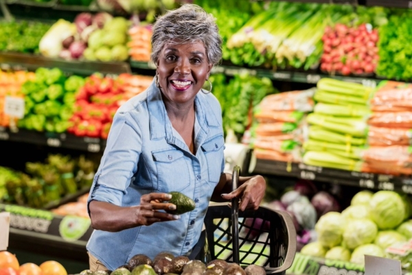 In a supermarket aisle surrounded by displays of fruit and vegetables a mature, smiling Black woman with short grey hair reaches for avocados.
