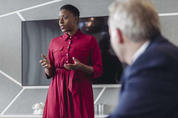A Black woman with short hair, red dress and her arms open addresses a meeting. Sat to her left is an unidentified white male in a blue suit. The walls are grey patterned and there is a screen behind her.
