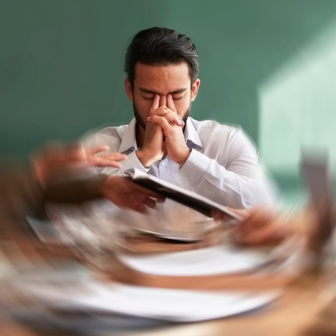 A concept of stress showing a business man holding his head in his hands while a motion blur shows colleagues around table handing him paperwork