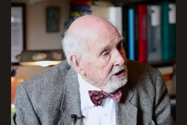 A colour portrait of Tim Travis-Healy looking off camera to his left. He is a white man with grey jacket and maroon bowtie.