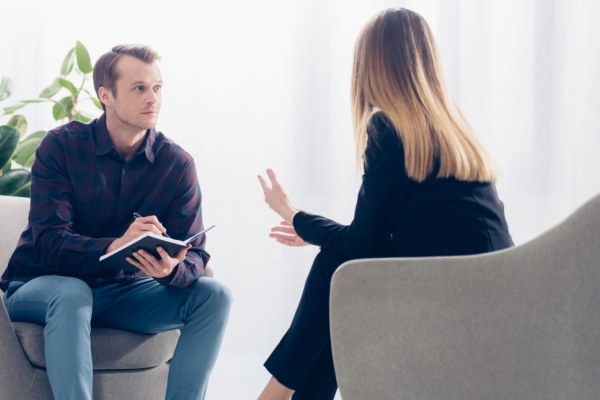 In a light filled room, a causally dressed white male journalist holds a notebook while interviewing smartly dressed white business woman with blond hair. The photo is taken from over her shoulder.
