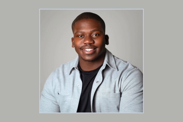 A portrait of Tariq Peters on a grey background. Tariq is a Black man with short hair and a light-grey shirt over a black t-shirt
