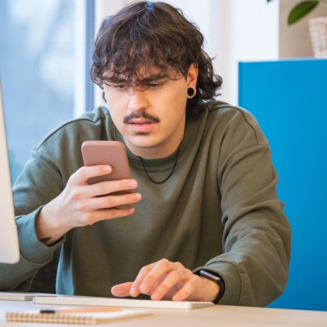 A young white man with dark hair and moustache grimaces as he stares at the mobile in his hand. He is sat in a front of a computer at a desk with a blue cabinet to the right of the image.