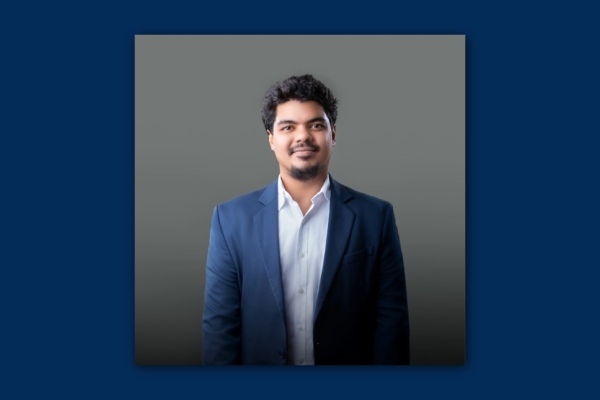 On a dark navy background a colour portrait of Thanzyl Thajudeen wearing a light blue shirt and navy suit. Thajudeen is an Asian man with short dark hair who smiles at the camera.