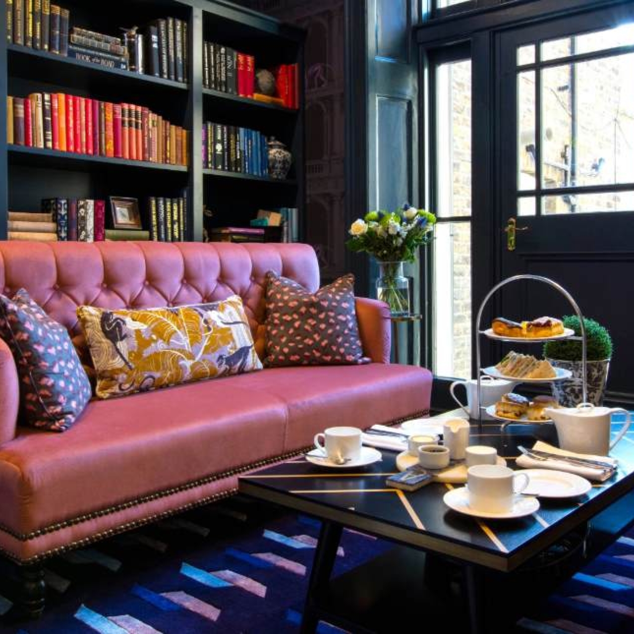 A dark blue room with a floor to ceiling bookcase, pink sofa and tan armchairs set around a coffee table with a traditional cream tea.