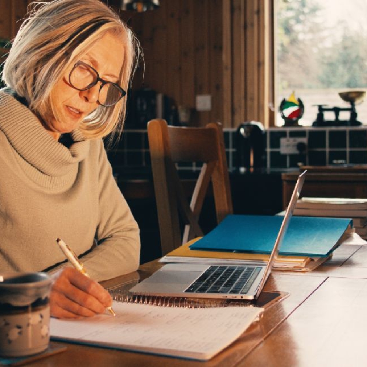 A middle aged white woman with a grey bob and beige jumper sat at a kitchen table writing in a notepad while surrounded by a laptop and paperwork.