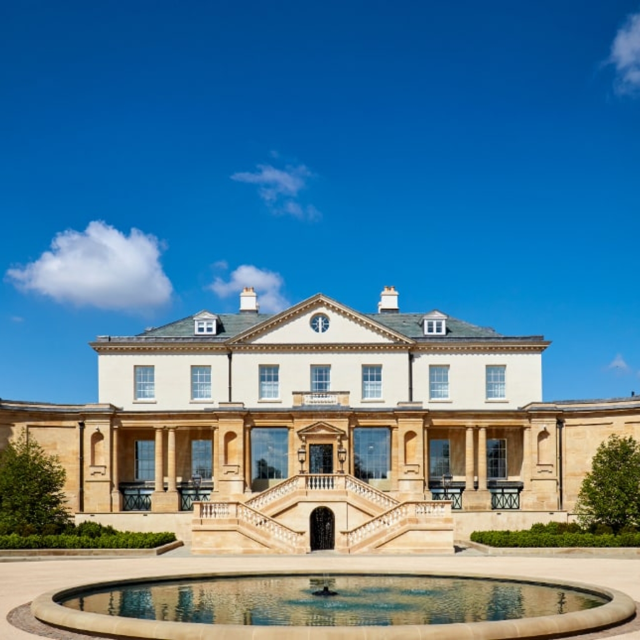A landscape shot of the outside of The Langley which is a pale building with a circular pond in front of it