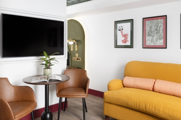 A picture of a hotel suite with white walls with pictures on them and a wall mounted TV, a table and brown chair and a yellow sofa
